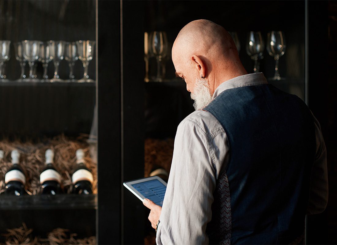 Contact - Rear View of a Senior Businessman Using a Tablet While Standing in Front of a Cabinet with Wine Glasses and Bottles